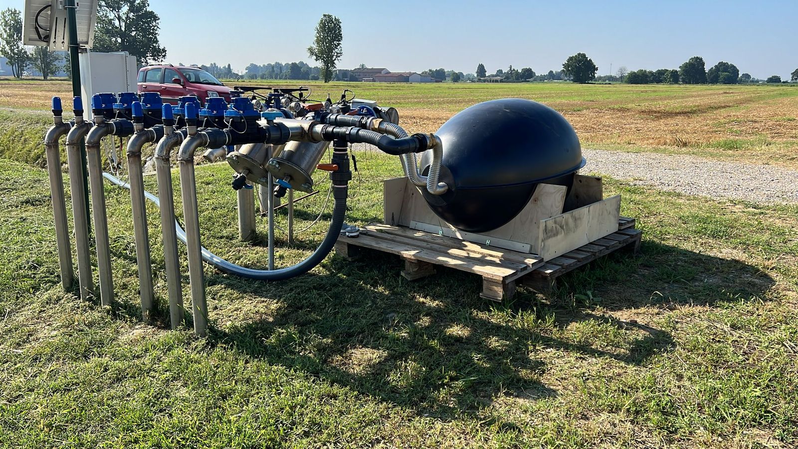 A black egg-shaped vessel on a wooden pallet, connected by hoses to an irrigation manifold of stainless steel pipes with blue valves, beside a field.