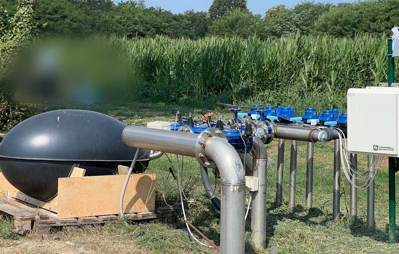 A dark egg-shaped vessel on a wooden frame, connected to stainless steel irrigation pipework with a row of blue valves; a grey electrical enclosure on a post at right and a stand of maize behind.