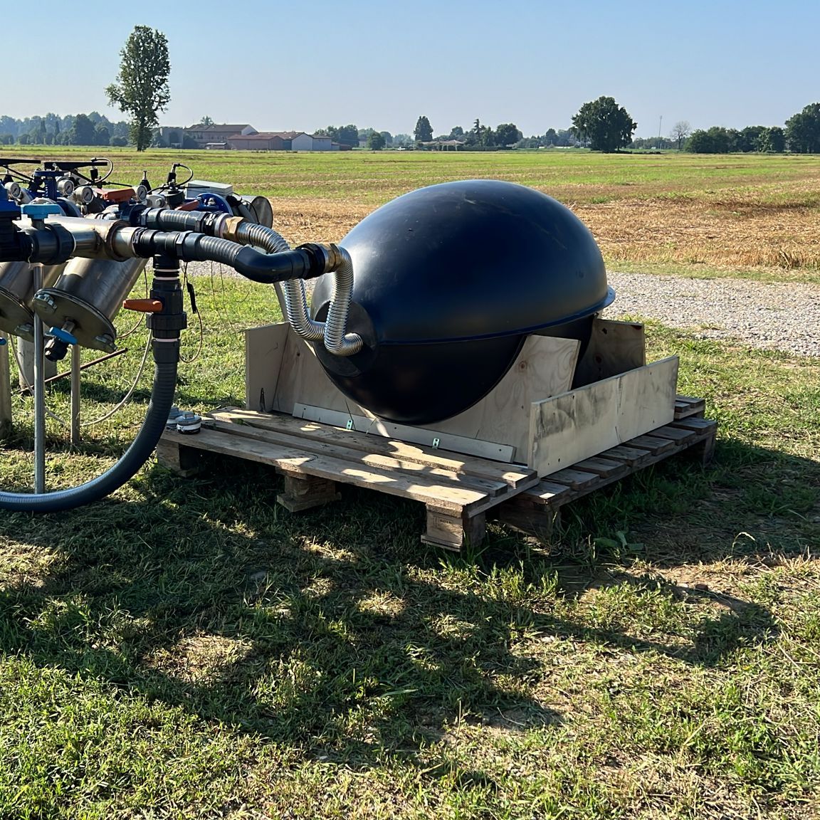 A black egg-shaped vessel on a wooden pallet, connected by hoses to the stainless steel filters and valves of an irrigation manifold, beside an open field.